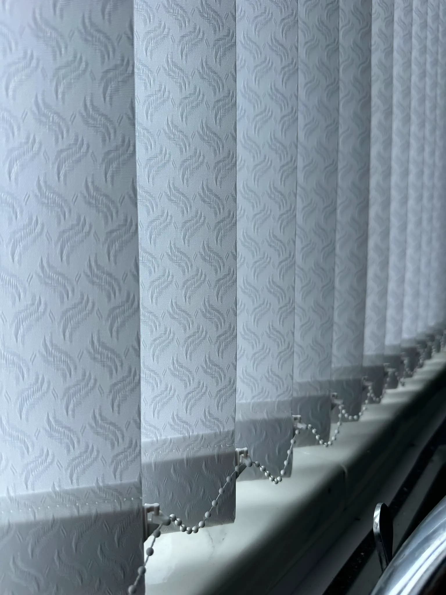 White leaf-textured vertical blind louvres with a chain-weight link along the sill
